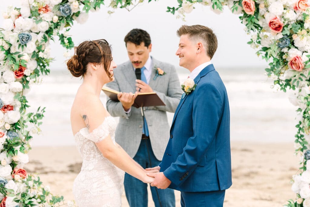 A couple stands hand in hand during their beach wedding ceremony, framed by an arch of flowers. One partner wears a lace gown, while the other is dressed in a blue suit. An officiant is present in the background, reading from a book.