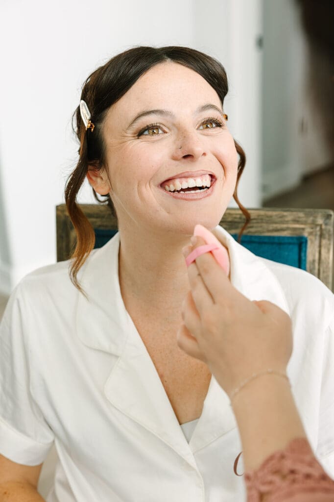 A bride smiling while receiving makeup application, sitting in a bright room with light-colored walls and furniture.