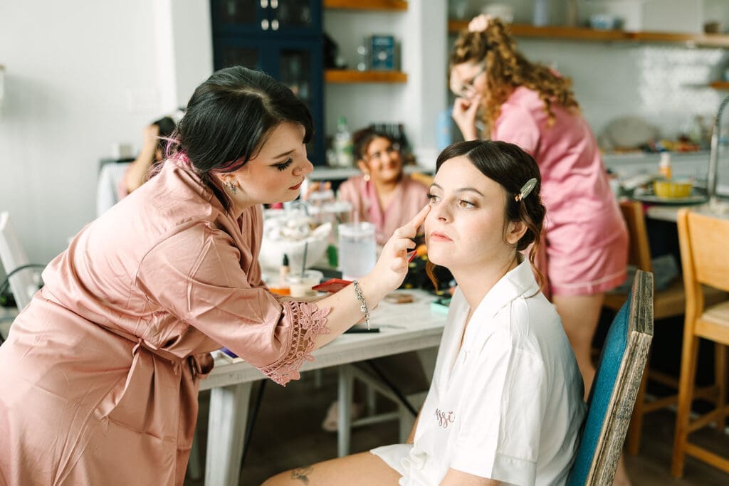 A stylist applies makeup to a bride in a cozy room filled with a group of friends, all dressed in soft robes, during a special occasion.