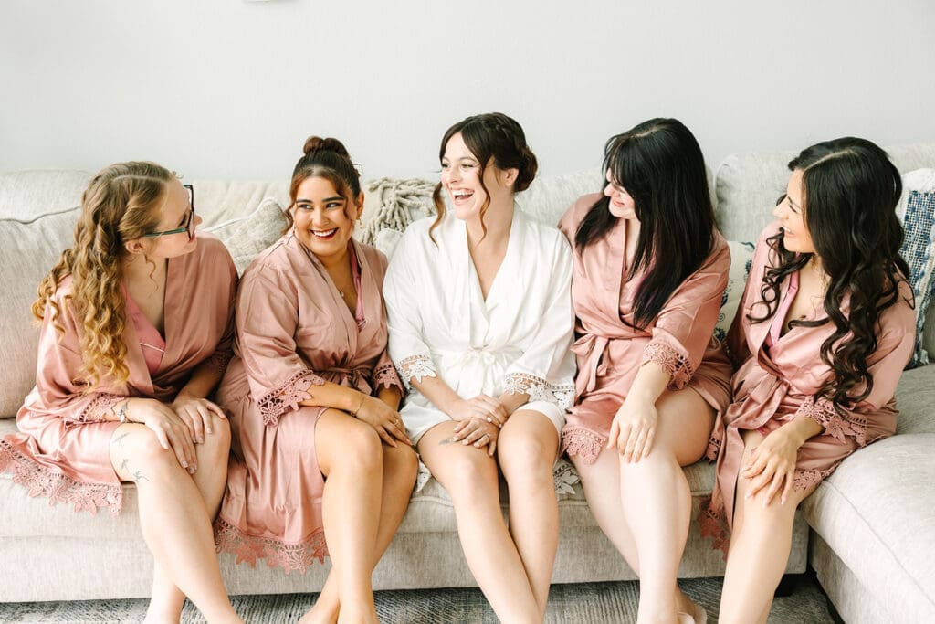Bride and bridesmaids in matching robes sitting on a couch, laughing and enjoying each other's company.