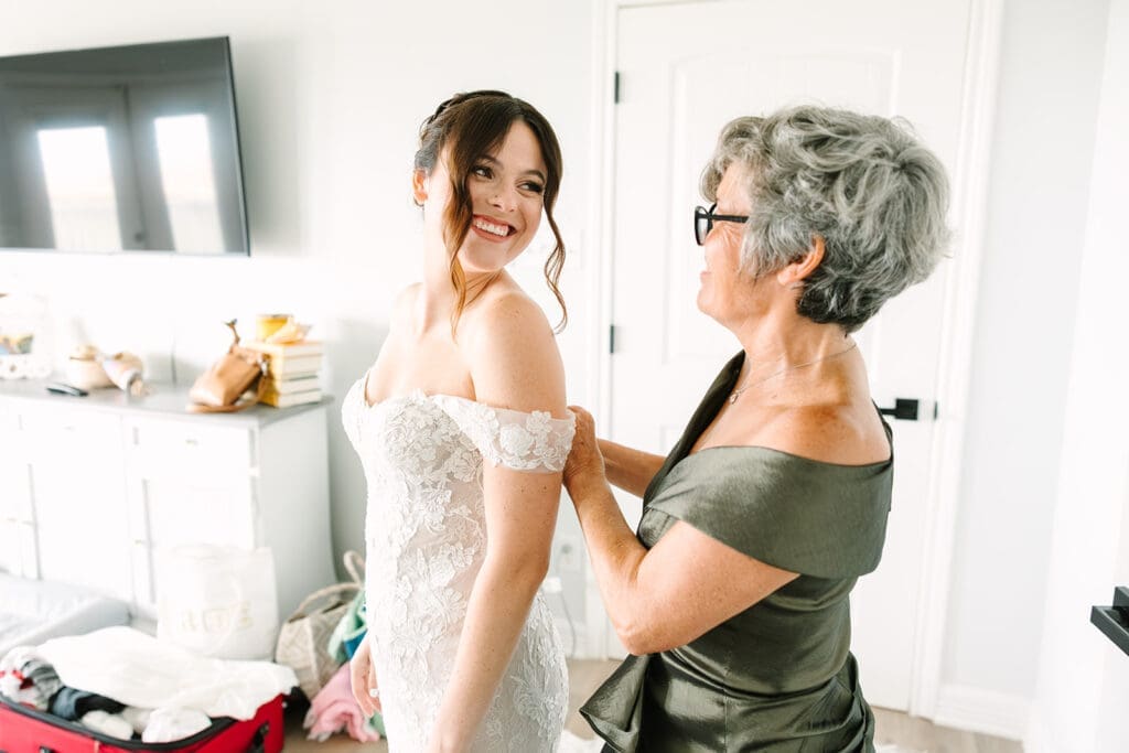 A bride in a wedding dress smiles while her mother helps her with the final adjustments in a bright, modern room.