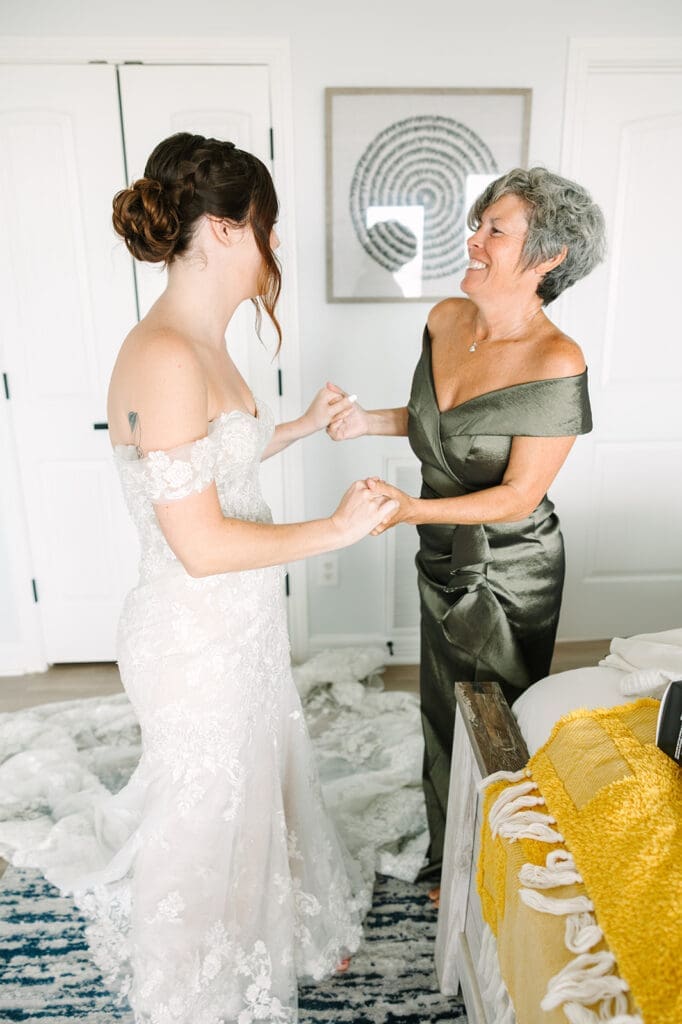 A bride in a lace wedding dress joyfully holds hands with her mother, who is wearing a green satin gown. They are both smiling and appear to be celebrating.