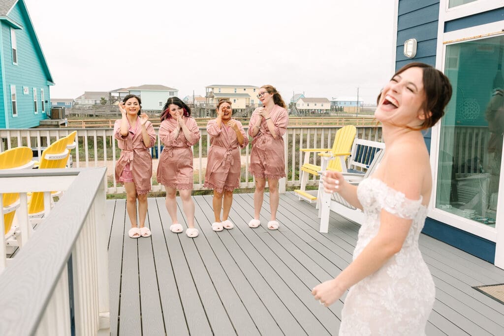 A bride in a white dress laughs joyfully while her bridesmaids, wearing pink robes, cheer and celebrate with excitement on a deck.