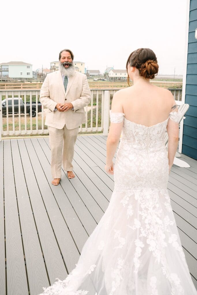 A father in a light suit stands on a deck, smiling and laughing as he looks at the bride in a lace wedding dress who is facing away from the camera.
