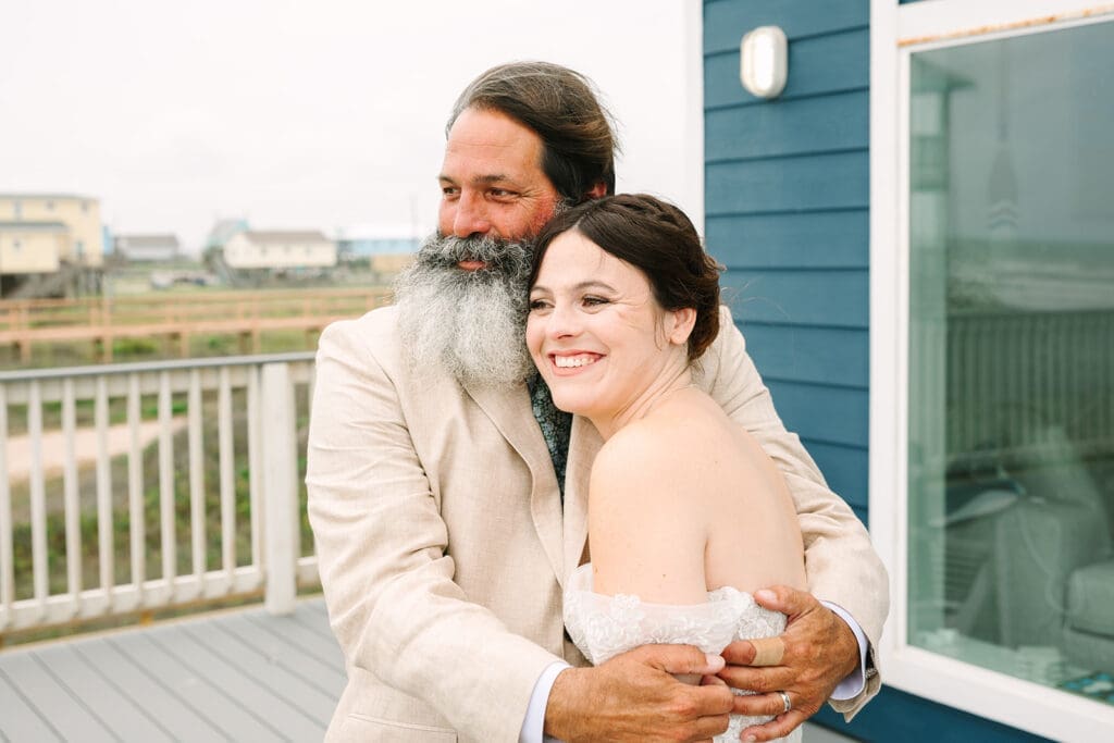 A smiling bride in a strapless wedding dress embraces her father with a beard, both in a coastal setting with houses in the background.