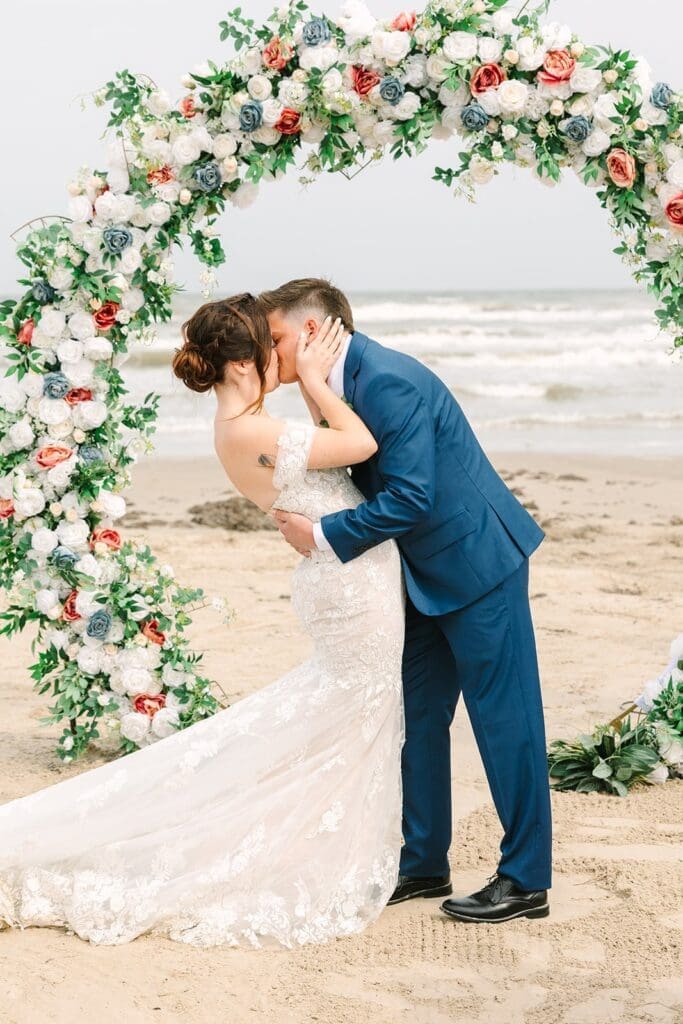 A bride and groom kissing under a floral arch at the beach, with the ocean in the background.