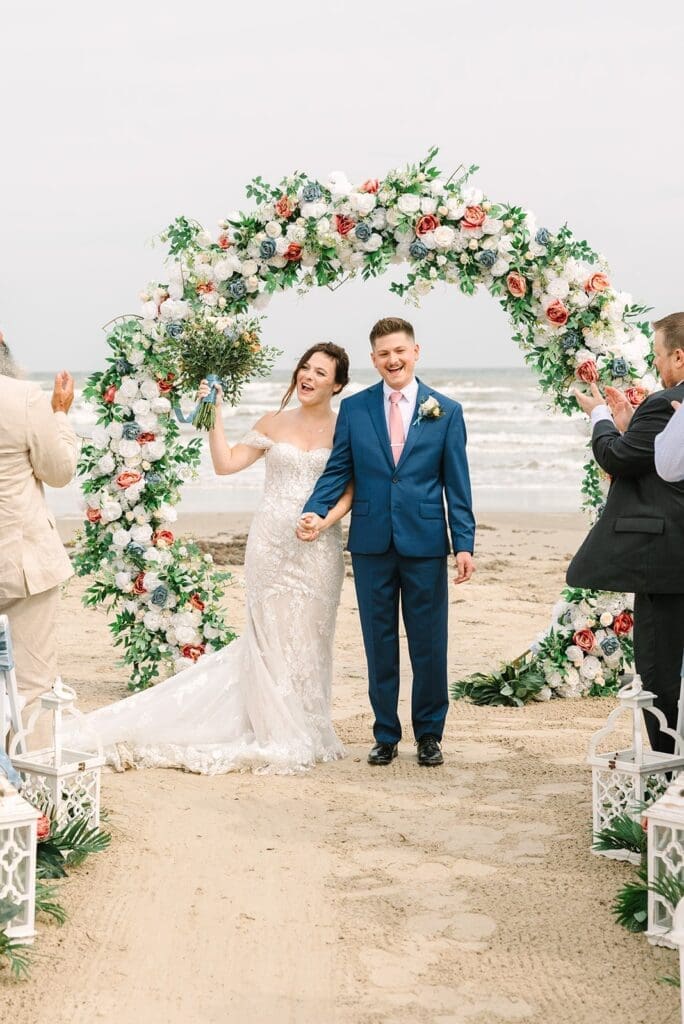 A bride and groom happily walk down the aisle on a beach, surrounded by floral decorations and guests. The bride holds a bouquet and wears an elegant wedding dress, while the groom is dressed in a blue suit with a pink tie.
