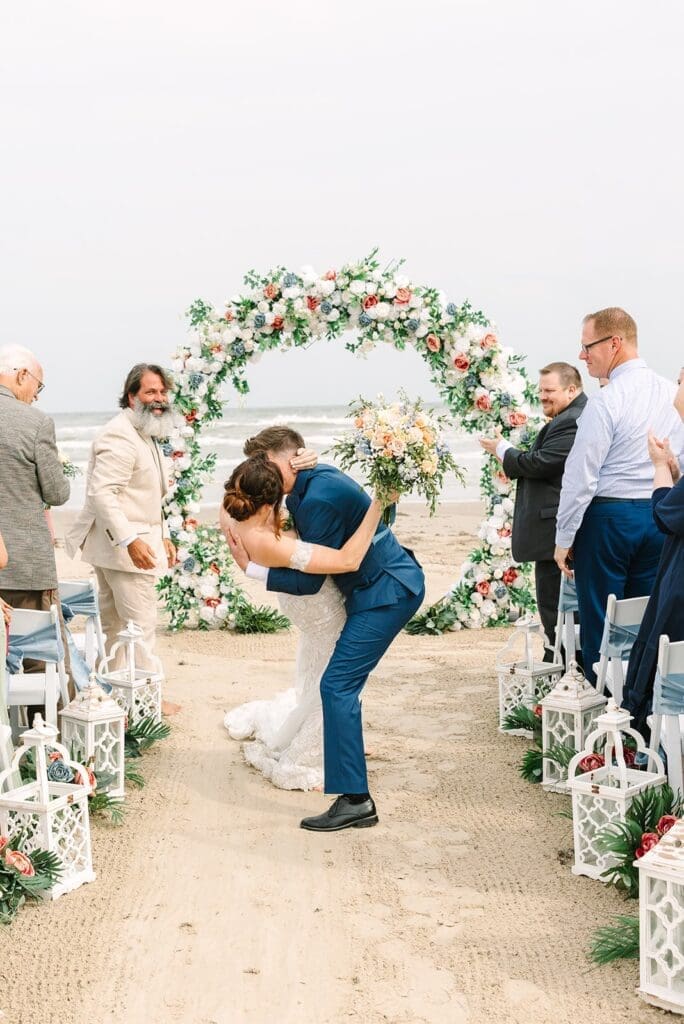 A newlywed couple sharing a kiss at their beach wedding ceremony, surrounded by floral decorations and smiling guests.