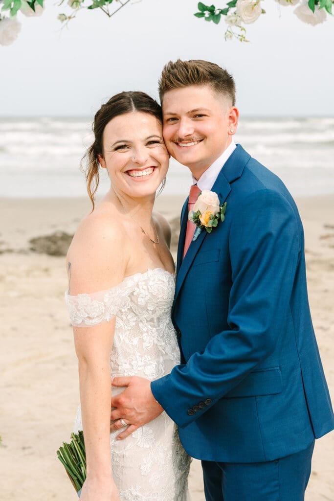 A happy couple posing together on a beach, with the woman in a lace wedding dress and the man in a blue suit, both smiling broadly. The background features a decorative floral arrangement and the ocean waves.