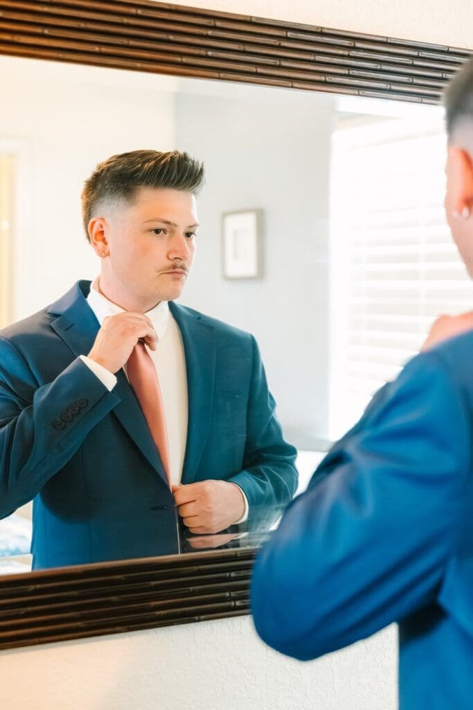 A man in a blue suit adjusts his pink tie while looking in a mirror.