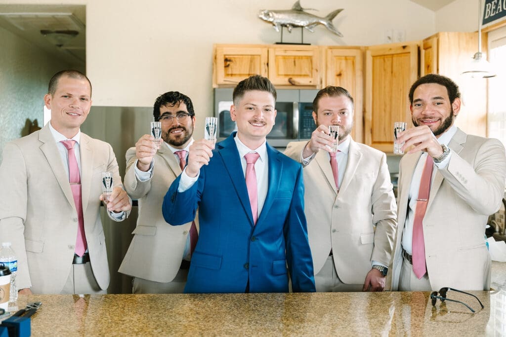 A groom and four men, dressed in light-colored suits with pink ties, are standing in a kitchen and holding up shot glasses in a toast. The man in the center is wearing a blue suit and is smiling.