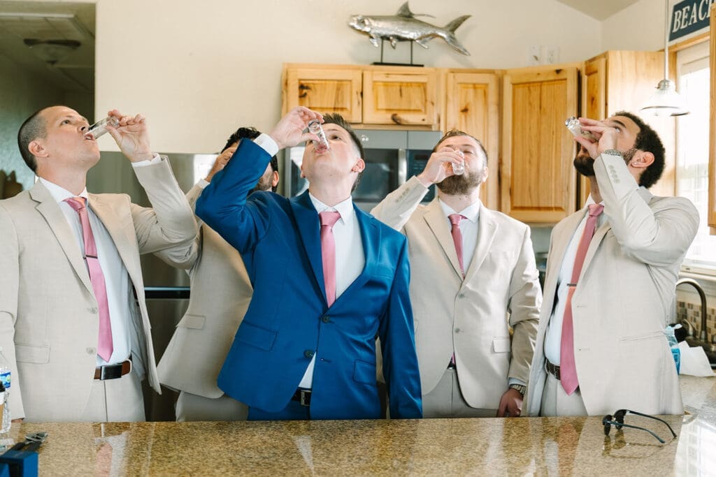 A groom and 4 men in light-colored suits are standing in a kitchen, raising shot glasses and preparing to take a drink. One man is wearing a blue suit. The kitchen has wooden cabinetry and kitchen appliances in the background.