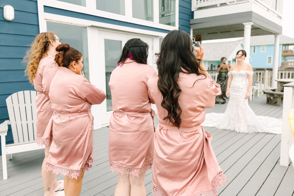 A bride in a white wedding dress is smiling while being photographed during first look with bridesmaids wearing pink satin robes.