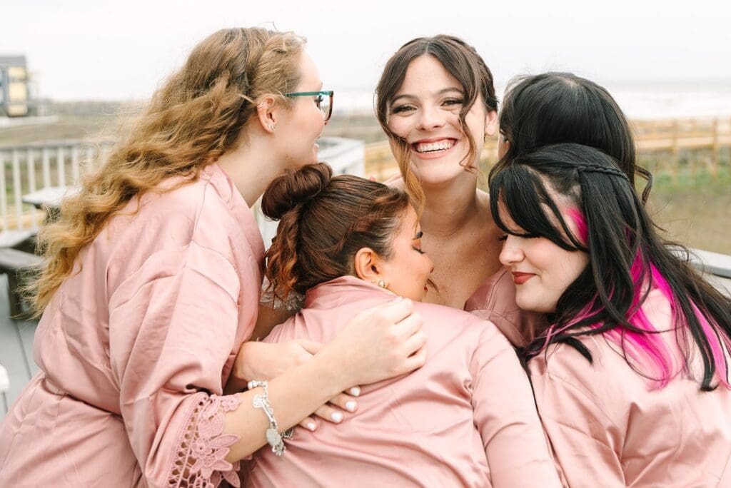 A group of five women wearing pink robes, joyfully embracing each other, with smiles and laughter, set against a background of a beach and ocean.